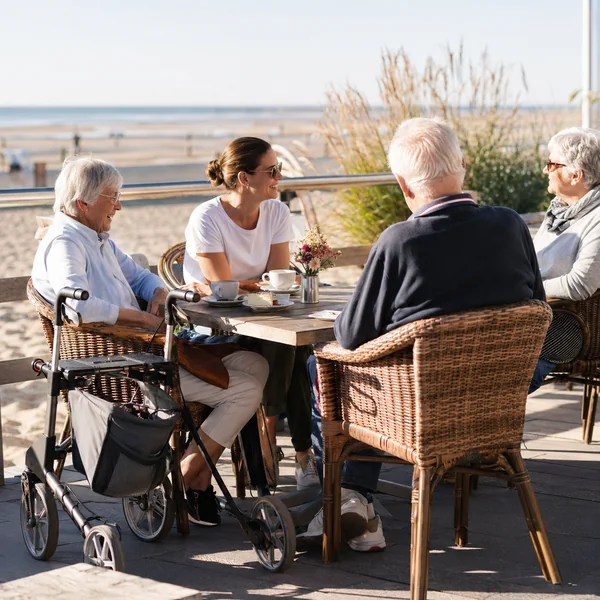 Reisen mit Demenz – gemeinsame Kaffeepause am Meer Ältere Menschen und eine jüngere Begleiterin bei einer Kaffeepause an der Strandpromenade, Rollator am Tisch