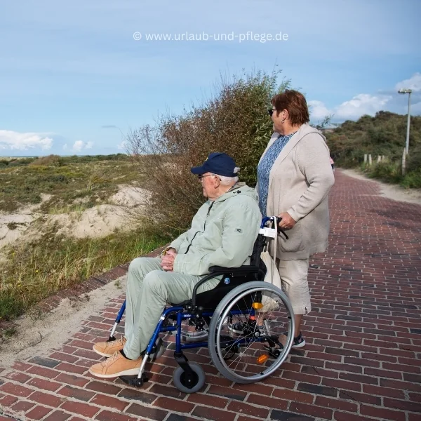 Betreute Reisen bei Demenz: Begleiterin und Senior im Rollstuhl blicken von einem gepflasterten Weg in die Dünenlandschaft an der Nordsee