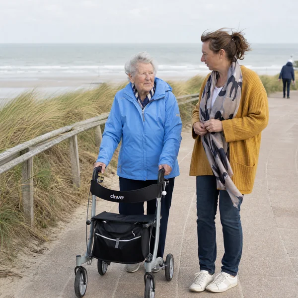Ältere Frau mit Rollator spaziert im betreuten Urlaub bei Demenz mit ihrer Reisebegleiterin auf der Promenade an der Nordsee