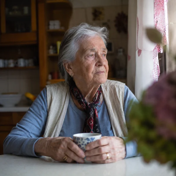 Nach der Demenz-Diagnose innehalten Ältere Frau sitzt nachdenklich am Fenster mit einer Tasse und blickt nach draußen