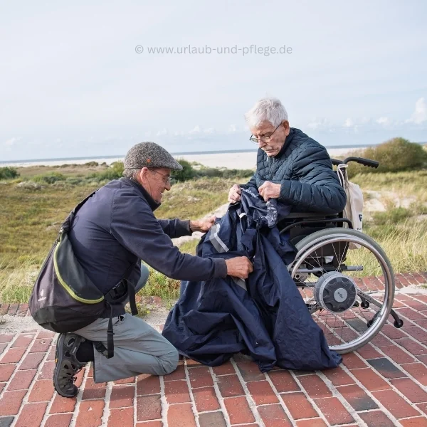 Urlaub mit Pflegegrad 5: Ehrenamtlicher Begleiter kniet vor einem Senior im Rollstuhl und hilft ihm am Strandweg mit dem Regencape