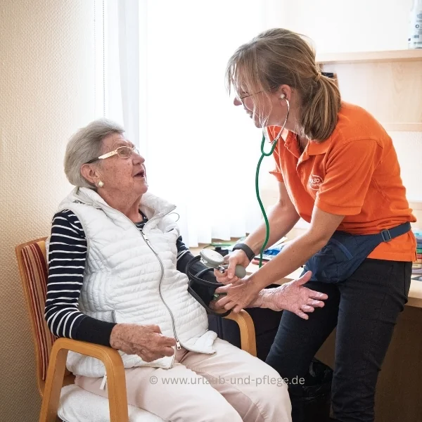 Urlaub mit Pflegegrad: Pflegefachkraft mit Stethoskop misst einer lachenden Seniorin im Hotelzimmer den Blutdruck