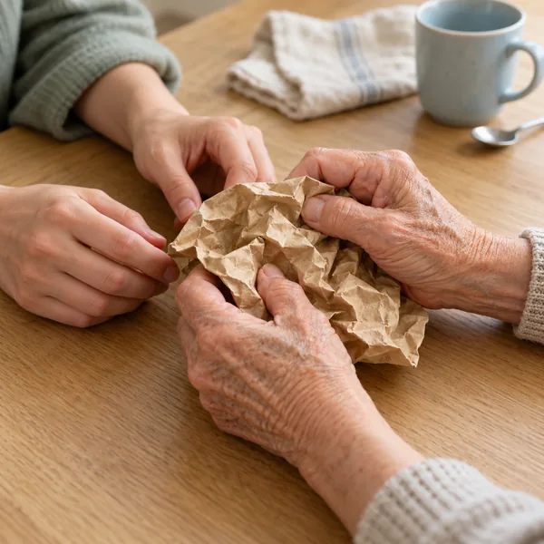 Ältere und jüngere Hände halten gemeinsam ein zerknittertes Stück braunes Papier auf einem Holztisch.